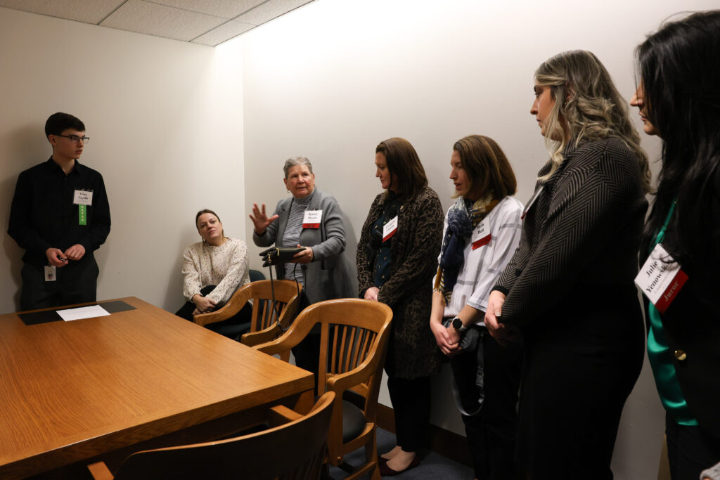 A group of volunteers jurors stand and sit in a small room to deliberate.